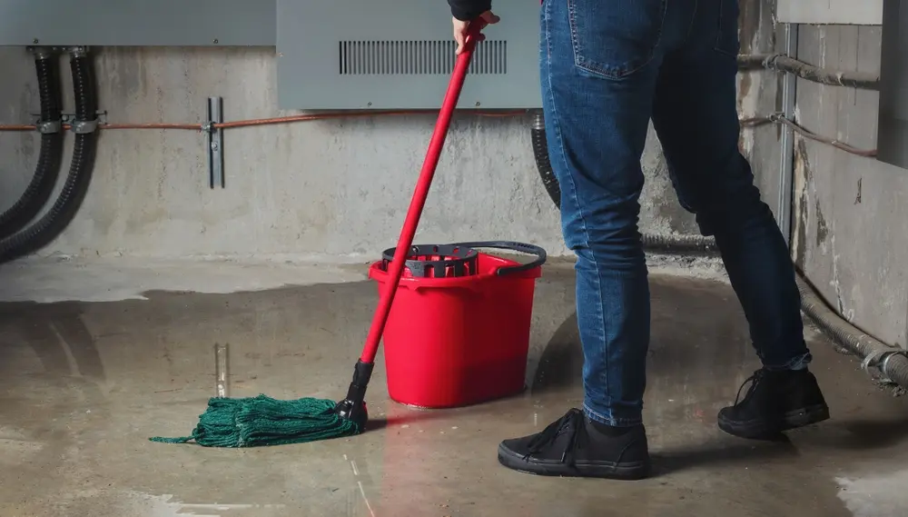 Woman mopping flood from water leaks in basement. Water damage from rain, snowmelt or pipe burst coming from multiple cracks and leaks in concrete wall and ceiling.